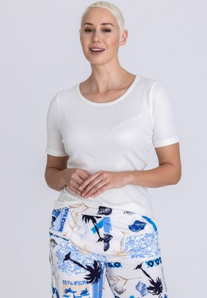 Femme aux cheveux blonds courts portant une chemise blanche à manches courtes et un pantalon blanc avec des motifs tropicaux de palmiers bleus et noirs et de coquillages.