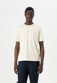 Cream short-sleeve t-shirt with a round neck and subtle logo on the chest, paired with dark pants and a smooth cotton texture.
