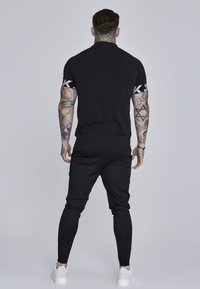 Čierny športový outfit so priliehavým krátkym rukávom a logom na rukávoch, doplnený o zúžené jogger nohavice. Biely tenisky dotvárajú vzhľad.
