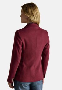 Kuldsete juustega naine, kes kannab keha poolest sobivat burgundi värvi bleiserit ja siniseid teksaseid, on näidatud seljaga tavalise tausta ees.