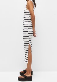 Femme portant une robe midi blanche sans manches avec des rayures horizontales noires et une fente sur le côté, assortie de sandales à plateforme noires, se tenant de côté.