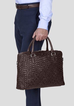 Brown woven leather briefcase with dual handles and silver hardware, worn by a person in business attire, displayed against a gray background.