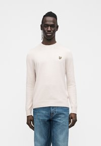 Ljusrosa stickad tröja med långa ärmar, rund halsringning och liten gul fjärilslogotyp på bröstet. Kombinerad med blå jeans i denim.