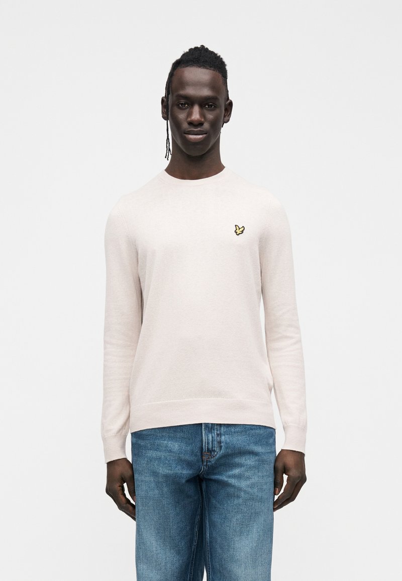 Ljusrosa stickad tröja med långa ärmar, rund halsringning och liten gul fjärilslogotyp på bröstet. Kombinerad med blå jeans i denim.