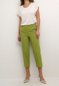 Vrouw in limoen groene high-waisted cropped broek, witte kortemouwtop en beige heeled mules, staand tegen een effen witte achtergrond.