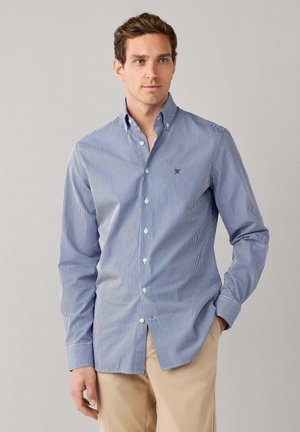 Uomo con camicia a righe blu e bianca abbottonata e pantaloni beige, in piedi con una mano in tasca davanti a uno sfondo semplice.