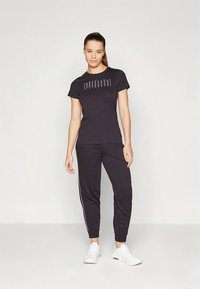 Juoda aptempi marškinėliai su spaustu tekstu, derinami su atitinkančiomis juodomis sportinėmis kelnėmis su šoninėmis juostelėmis. Modelis dėvi baltus sportinius batelius.