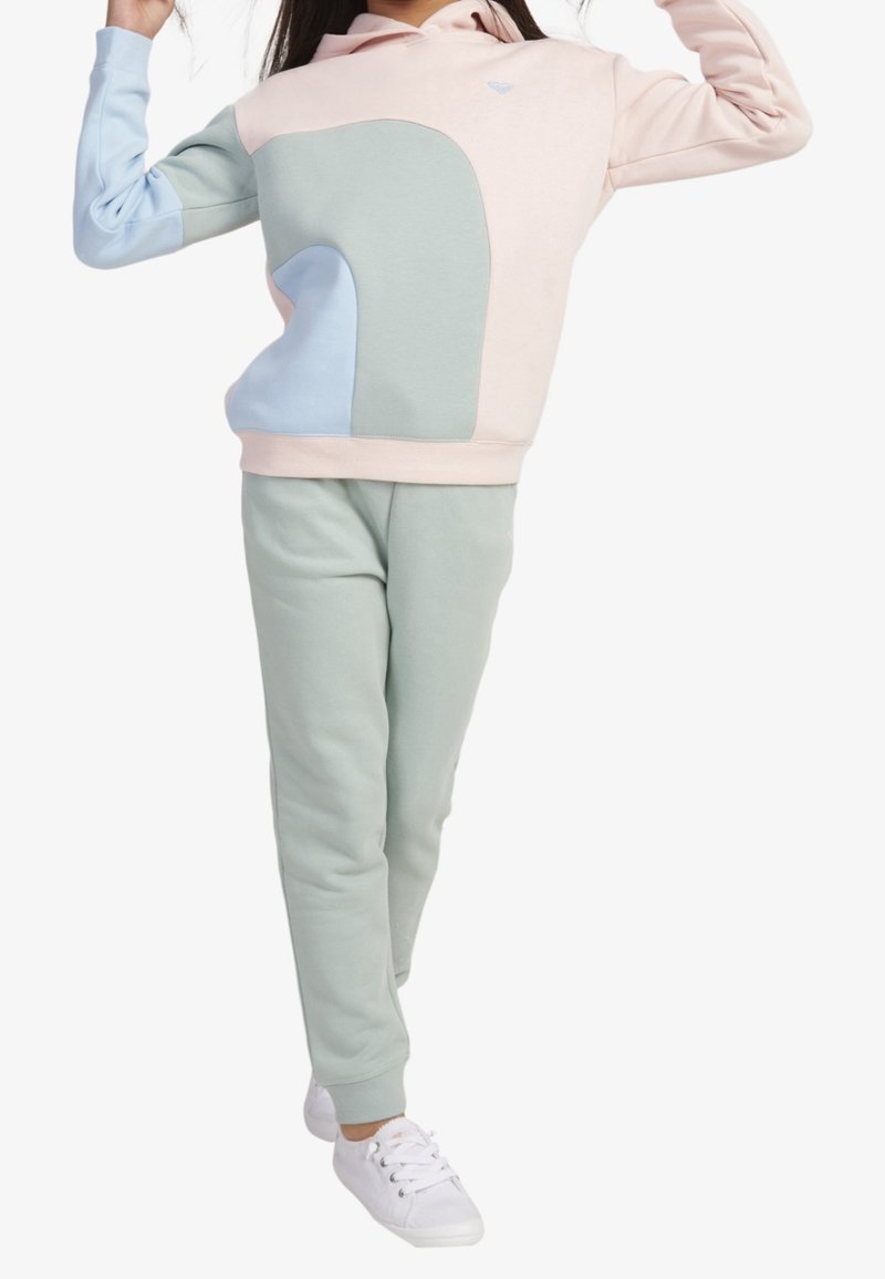 Fille portant un sweat à capuche color-block pastel rose, bleu et vert avec un pantalon de survêtement vert assorti et des baskets blanches, posant les bras levés.