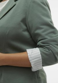 Zelený blejzr s hladkou texturou, zdobený bílým proužkovaným manžetou. Materiál je lehký, s dobře padnoucím střihem a klasickými klopy.