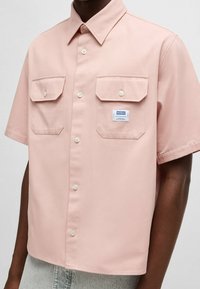 Chemise à manches courtes rose à boutons, avec deux poches poitrine, boutons blancs et une étiquette bleue portant "Hugo" sur le côté gauche. Texture lisse.
