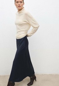 Un pull à col roulé beige assorti à une jupe maxi marine. La tenue présente des textures lisses et des silhouettes ajustées, complétées par des bottes à talons marron.