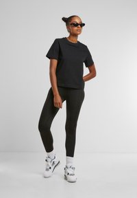 Zwart katoenen T-shirt met korte mouwen, gecombineerd met zwarte leggings. Het model draagt witte sneakers met teal accenten en zwarte zonnebril.