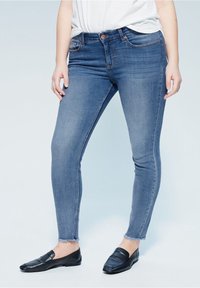 Jeans skinny de mezclilla azul con un diseño ajustado, dobladillo deshilachado y un desgaste ligero en la parte frontal. Combinados con mocasines negros para un look casual.