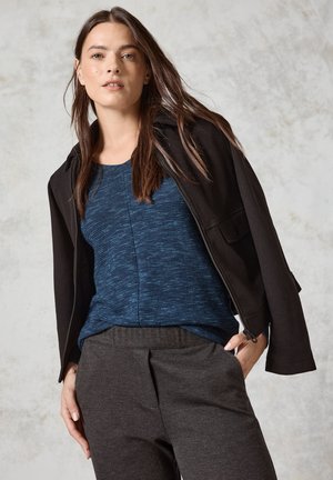 Veste zippée noire sur un haut texturé bleu, associée à un pantalon gris fuselé. Cheveux naturels tombent librement autour des épaules.