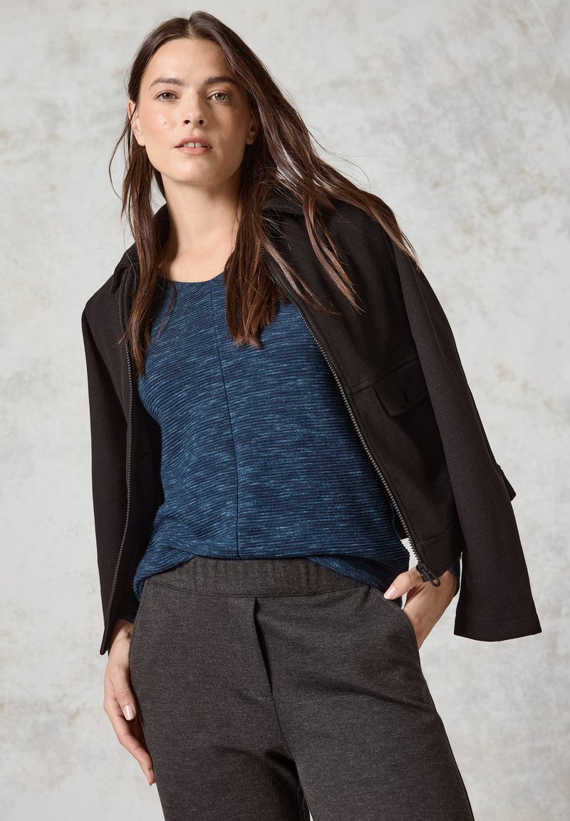 Svart zip-up jacka över en blå texturerad topp, i kombination med grå tapered byxor. Det naturliga håret faller löst runt axlarna.