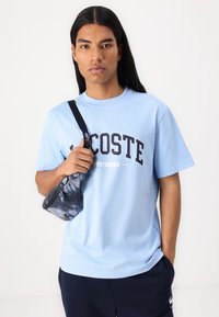 Světle modré bavlněné tričko s logem "LACOSTE" v tmavě modré barvě, kulatý výstřih, krátké rukávy. Drží černou a modrou vzorovanou tašku.