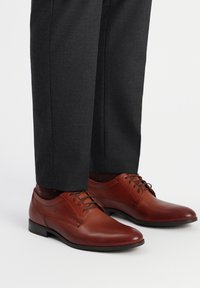 Zapatos de vestir de cuero marrón con textura suave, diseño de cordones, punta redondeada y sutiles detalles laterales, combinados con pantalones gris oscuro.