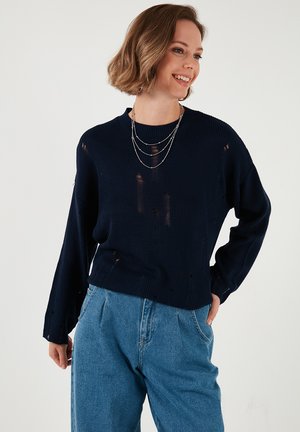 Navy knitted sweater with distressed detailing, long sleeves, and a crew neckline. Paired with high-waisted denim trousers.