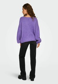 Maglione oversize viola lavorato a maglia con maniche a palloncino, indossato con jeans dritti neri e stivali chunky neri. Dettaglio in filato testurizzato.