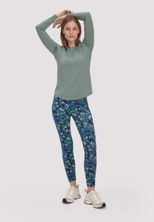 Vrouw die stretcht met opgetrokken armen, gekleed in een groene top met lange mouwen, blauwe legging met patroon en witte sportschoenen, tegen een effen achtergrond.