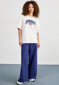 Krämfärgad oversize T-shirt med marinblå grafisk logga, kombinerad med vida marinblå byxor med sidoremmar. Vita sneakers fullbordar outfiten.
