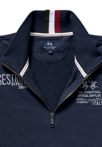 Mörkblå pikétröja med halvdragkedja vid kragen, texturerat tyg, vita och färgade randiga detaljer samt broderade logotypdetaljer. Storlek M.