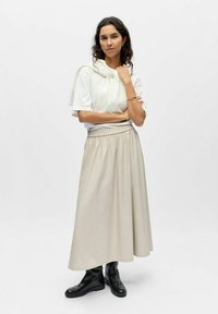 Femme aux cheveux bouclés portant un t-shirt blanc, une longue jupe beige, des bottes noires et un pull blanc posé sur les épaules, sur un fond uni.