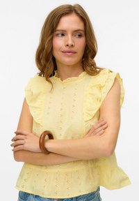 Blusa di cotone gialla con maniche a strascico, caratterizzata da fantasie ad occhiello e un colletto scallop, abbinata a un bracciale di legno al polso.