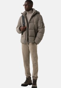 Szary puchowy płaszcz o teksturowanym wykończeniu i przypiętym kapturze, zestawiony z jasnym golfem, beżowymi spodniami i ciemnymi sznurowanymi butami.