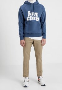 Blå hoodie med vit text "fan club", beige byxor och vita sneakers med marinblå och röda detaljer, stående på en neutral bakgrund.