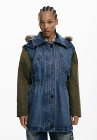 Denim a nylon parka so zelenými rukávmi, kapucňou s pravou kožušinou, zapínaním na gombíky a vreckami. Má voľný strih a kontrastné materiály.