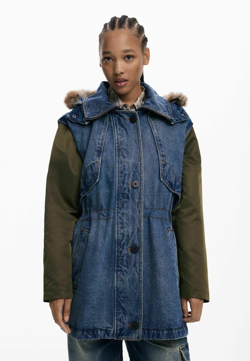 Denim a nylon parka so zelenými rukávmi, kapucňou s pravou kožušinou, zapínaním na gombíky a vreckami. Má voľný strih a kontrastné materiály.