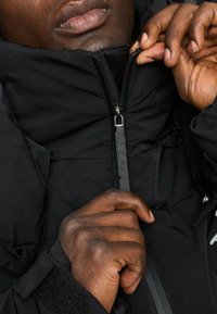 Giacca nera isolata con colletto alto e zip centrale. Presenta un tirazip in gomma e polsini in velcro. Tessuto liscio.