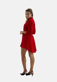 Robe rouge à manches longues avec poignets à boutons, présentant une coupe décontractée et un ourlet asymétrique. Portée avec des chaussures à talons hauts noires.