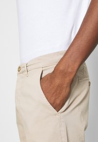 Pantalones de algodón beige con diseño de corte recto, que cuentan con bolsillos laterales y un detalle de botón en la cintura. Combinados con una camiseta blanca.