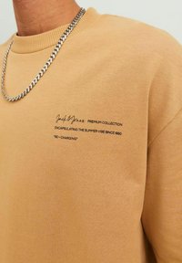 Beige pamut pulóver bordázott gallérral, elöl fekete szövegdesignnal, amely tartalmazza a márkanevet és a szlogent. Ezüst lánc nyaklánc látható.