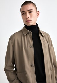 Brun button-up overtøj med en tekstureret finish, parret med en tætsiddende sort turtleneck. Har en krave og forlommer.