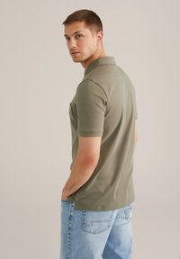 Camiseta polo de color verde oliva con manga corta y un bolsillo en el pecho, combinada con unos vaqueros de denim azul claro. Diseño simple, sin patrones ni gráficos.