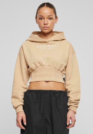 Beige, kort hoodie med snøring i hetten og ribbestrikket kant. Har hvit logo på fronten. Kombinert med løse sorte bukser, som fremhever kontrasten.