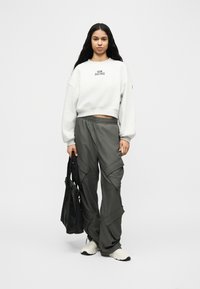 Νεαρή γυναίκα με μακριά σκούρα μαλλιά φοράει λευκό φούτερ New Balance, γκρι παντελόνι cargo, λευκά αθλητικά παπούτσια, κρατώντας μαύρη τσάντα ώμου, στέκεται μπροστά σε γκρι φόντο.