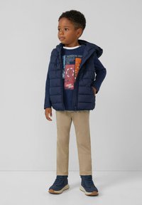 Marineblauwe gewatteerde vest met capuchon over een donkerblauwe grafische t-shirt, beige broek en marineblauwe sneakers met gestructureerde zolen. Eenvoudig ontwerp en kleuren.