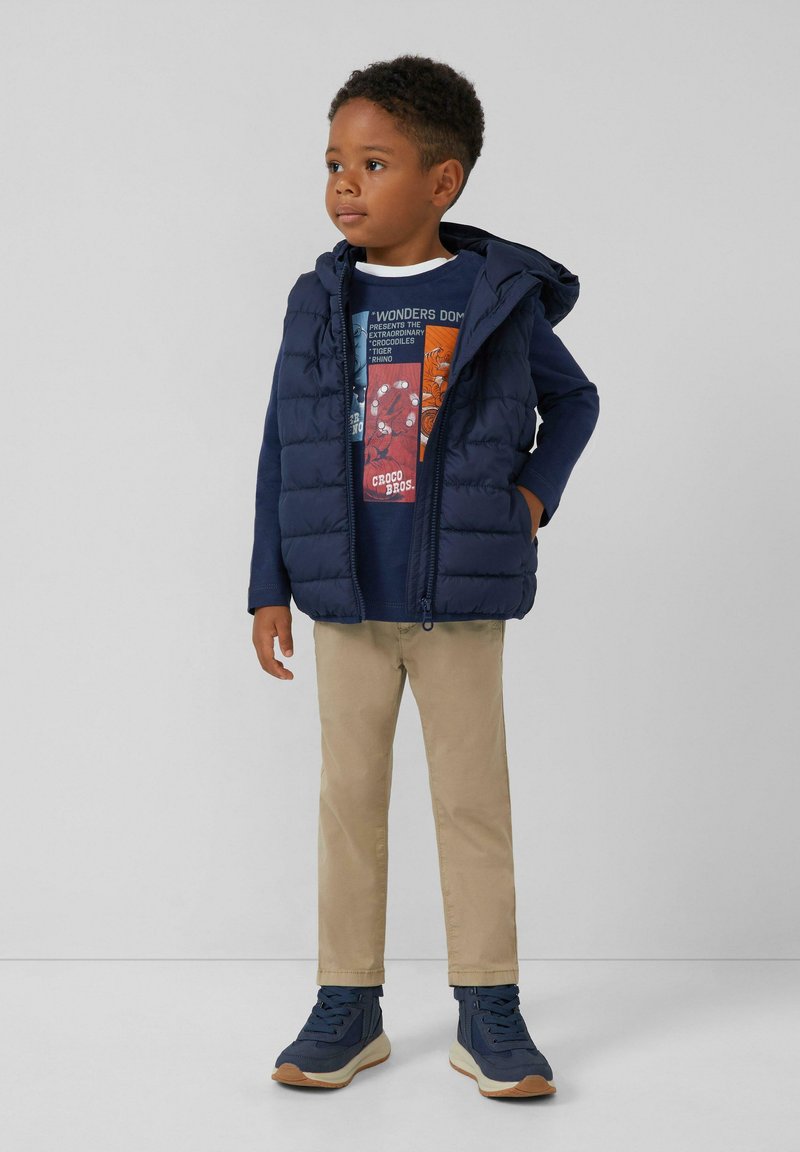 Marineblauwe gewatteerde vest met capuchon over een donkerblauwe grafische t-shirt, beige broek en marineblauwe sneakers met gestructureerde zolen. Eenvoudig ontwerp en kleuren.