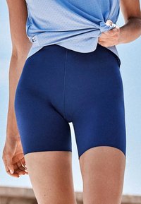Personne portant un short bleu marine ajusté et un haut perforé bleu clair, soulevant l'ourlet du haut d'une main.