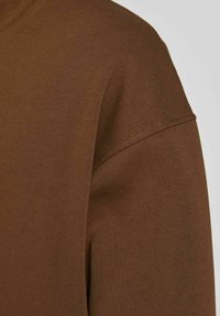 Tela de camiseta de algodón marrón exhibida, con una textura suave, mangas cortas y un cuello redondo con detalle de costura visible.