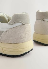 Grijze suède en witte mesh sneakers met een ronde hak, textuuraccenten en een contrasterende gele tussenzool en zwarte buitenzool.