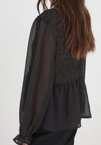 Blusa nera trasparente con pois bianchi, presenta maniche a sbuffo, vita arricciata e una silhouette fluida. Tessuto morbido e leggero.