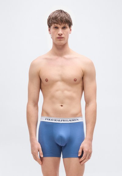 Μπλε ανδρικά μποξεράκια με λευκή ταινία που φέρει το μήνυμα "POLO RALPH LAUREN" σε μαύρο χρώμα. Μαλακή υφή και εφαρμοστή σχεδίαση. Το μοντέλο στέκεται με τα χέρια δίπλα στο σώμα.