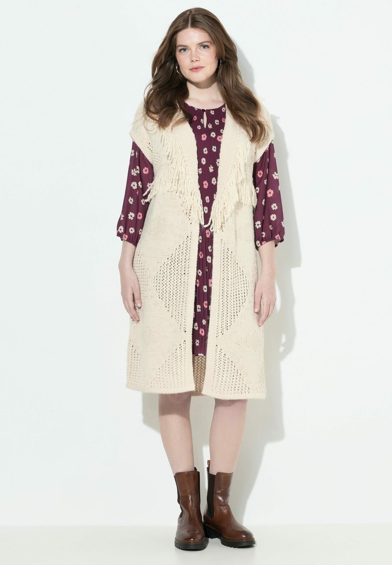 Häkelweste in Creme mit Fransen über einem weinroten Blumenkleid. Braune Knöchelstiefel. Gemustertes Design und offene Vorderseite.