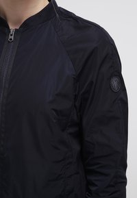 Närbild av en person som bär en svart zip-up bomberjacka med ribbat krage och en rund patch på övre ärmen.
