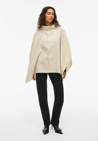 Beige uld poncho med høj krave og sømløst design, parret med sorte fløjl bukser og spidse sorte højhælede sko.